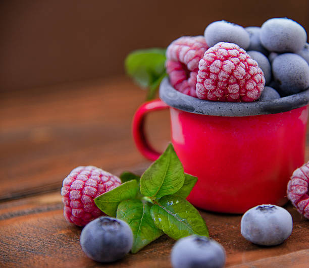 Close-up of frozen raspberries and blueberries in a red enamel mug on wooden background. Rustic winter concept with frosty berries and vibrant natural colors.
