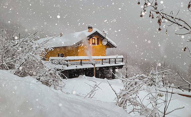 A yellow mountain cabin surrounded by heavy snowfall and blanketed trees during a serene winter storm. Smoke rises from the chimney, adding warmth to the cold, peaceful landscape.