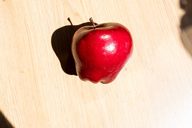 Vibrant, shiny red apple sits on a light wooden table, casting a distinct shadow under the bright sunlight, creating a simple yet appealing image of freshness and natural goodness