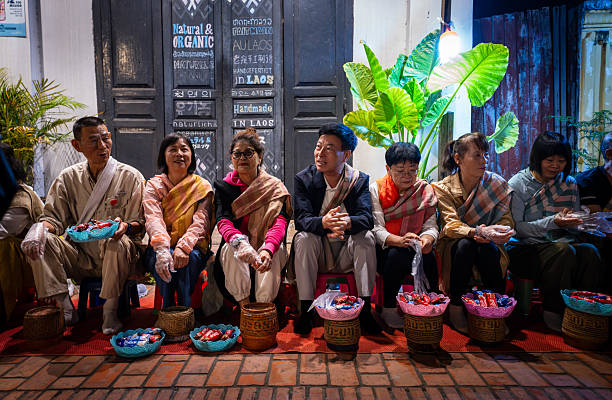 Luang Prabang, Laos - November 9, 2024：The warm lights of a Wat in Luang Prabang, Laos, provide a backdrop for a group of people with baskets, possibly waiting for or participating in an evening ritual or ceremony.