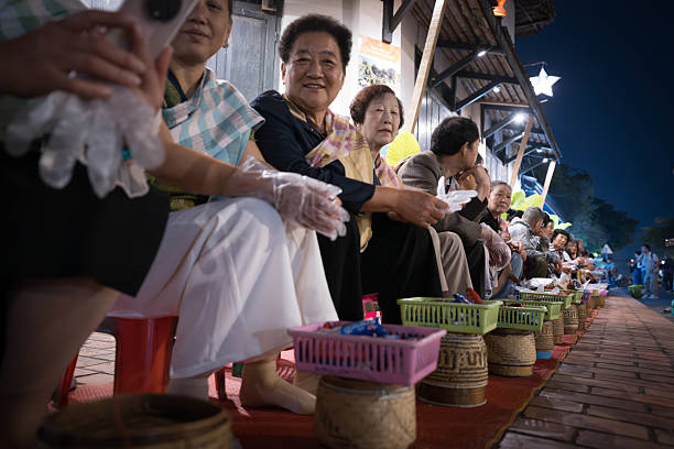 Luang Prabang, Laos - November 9, 2024：The warm lights of a Wat in Luang Prabang, Laos, provide a backdrop for a group of people with baskets, possibly waiting for or participating in an evening ritual or ceremony.