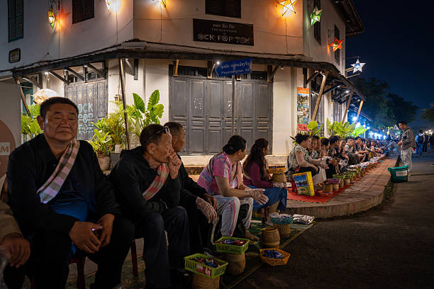 Luang Prabang, Laos - November 9, 2024：The warm lights of a Wat in Luang Prabang, Laos, provide a backdrop for a group of people with baskets, possibly waiting for or participating in an evening ritual or ceremony.