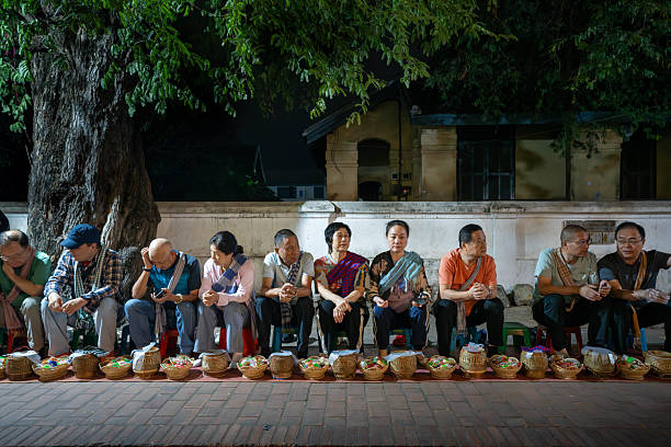 Luang Prabang, Laos - November 9, 2024：The warm lights of a Wat in Luang Prabang, Laos, provide a backdrop for a group of people with baskets, possibly waiting for or participating in an evening ritual or ceremony.