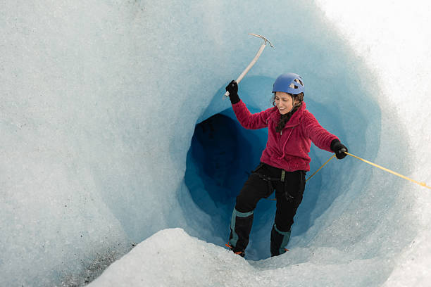 Female mountaineer smiles while carefully entering a glacial cave, using an ice axe and rope for safety