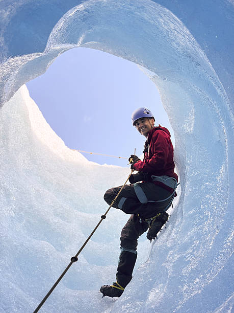 Low angle view capturing a mountaineer smiling at the camera while ascending a moulin, surrounded by stunning blue ice and rugged glacier terrain