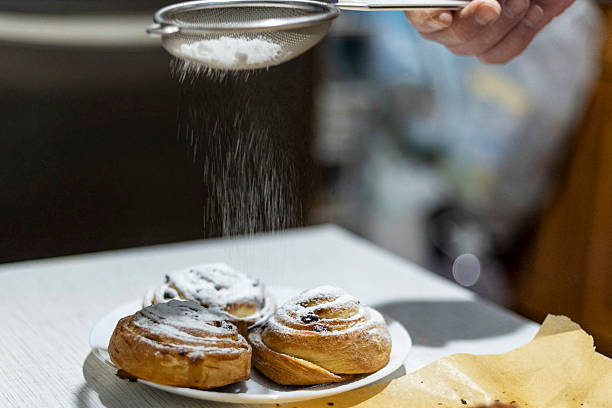 A hand dusts powdered sugar through a sieve over freshly baked cinnamon rolls in a kitchen setting. The image captures the process of adding a sweet finishing touch to the pastries.