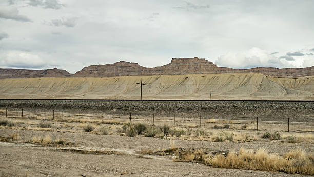 A desolate desert scene featuring a single set of train tracks running past eroded sandstone cliffs under a cloudy sky, with sparse vegetation and a leaning utility pole