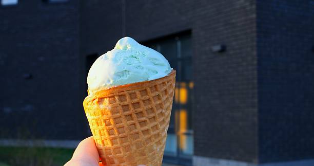 istock Melting pistachio ice cream dripping along waffle cone, hand held against blurred black urban building with illuminated windows during warm sunset moments 2213158763