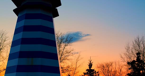 Decorative blue and white striped lighthouse isolated on white background, set against a backdrop of a beautiful sunset with orange, pink, and blue hues blending seamlessly