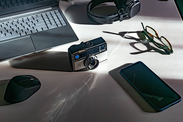 Madrid. Spain. A vintage camera sits on a white desk alongside modern technology such as a laptop, smartphone, headphones, and glasses, creating a striking contrast between past and present.