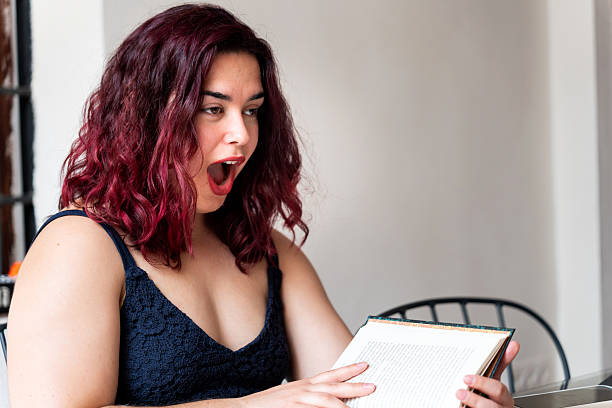Body positive young woman with colorful hair reading a book and looking surprised by the plot twist