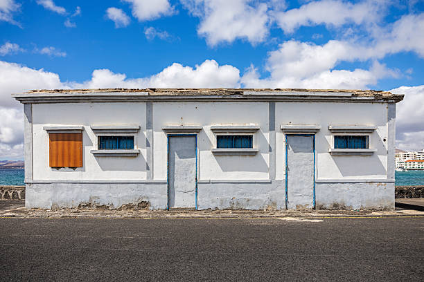 Arrecife, Lanzarote, Canary Islands, Spain - March 11st 2025:  A small, weathered building stands on a Spanish harbor, its white walls accented with blue trim. The structure features two doors and three windows, some with protective bars and one covered by a wooden shutter. Signs of aging, including peeling paint and worn surfaces, hint at its long-standing presence by the waterfront. Above, a bright blue sky with scattered clouds enhances the contrast between the rustic building and the open expanse. In the background, the sea and distant buildings complete the maritime setting, offering a glimpse into the character of coastal life.