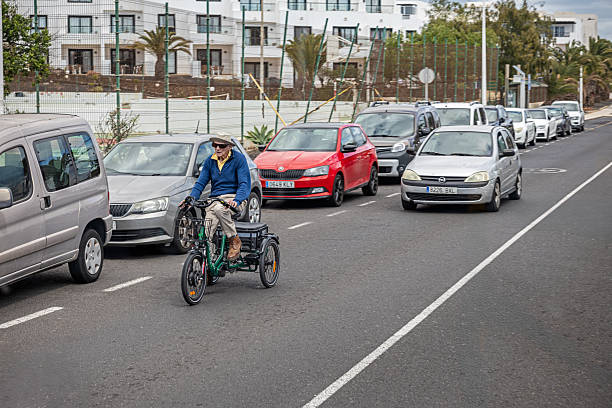 Arrecife, Lanzarote, Canary Islands, Spain - March 12th 2025:  This street scene captures an elderly man navigating city traffic on a three-wheeled bicycle. Dressed in a blue sweater, beige pants, and a beige hat, he pedals steadily along the road, his tricycle equipped with a front basket and a rear storage compartment. Surrounding him are parked cars, including a red vehicle with a visible license plate, emphasizing the contrast between traditional cycling and modern transportation. In the background, contemporary buildings and a fenced area with palm trees frame the urban setting, adding depth to the composition. This image highlights themes of independence, sustainable mobility, and the coexistence of different modes of transport in a bustling city.