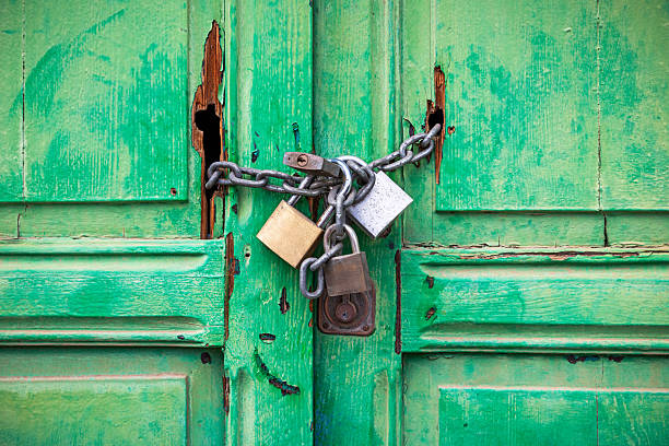 This compelling image captures a weathered green wooden door in Arrecife, Lanzarote, tightly secured with a chain and multiple padlocks. The door’s peeling paint and visible cracks hint at years of exposure to the coastal climate, while the intertwined chains and varied padlocks reinforce the sense of security and restricted access. The contrast between the aged door and the heavy-duty locks suggests a story of protection, abandonment, or secrecy. The surrounding urban textures add depth to the scene, making it a striking representation of resilience and mystery in Lanzarote’s capital.