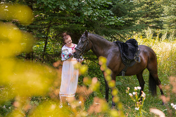 A woman dressed in a traditional outfit stands lovingly beside a horse in a tranquil forest. Soft sunlight filters through the trees, creating a peaceful atmosphere.