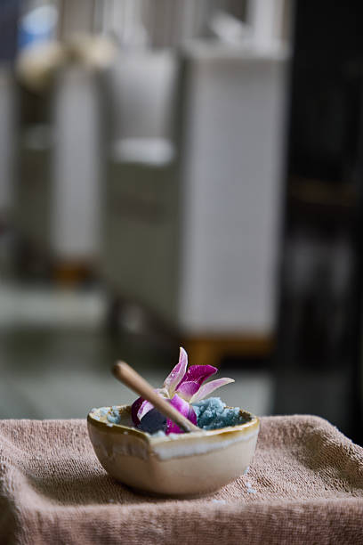 A decorative ceramic bowl containing blue salt and a pink flower placed on a brown cloth, evoking relaxation and spa atmosphere with blurred background chairs creating a tranquil setting.