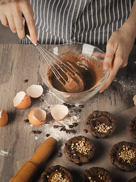 istock Female hands beat with a whisk dough into a bowl next to muffins, eggshell and rolling pin on on a table, faceless baker in striped apron, vertical image. 2213185171