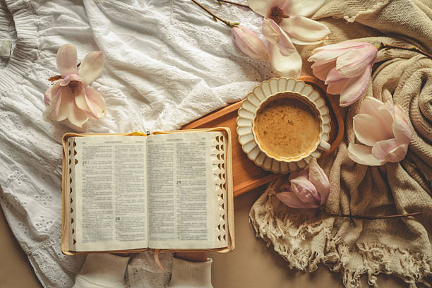 Person holding a cup of coffee next to an open Bible, surrounded by blooming magnolia flowers and soft fabrics, evoking a peaceful and reflective spring morning