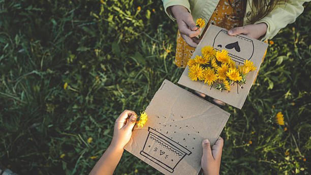 Two children creating flower crafts with fresh dandelions on cardboard, enjoying springtime nature and creative outdoor activity in a grassy field.