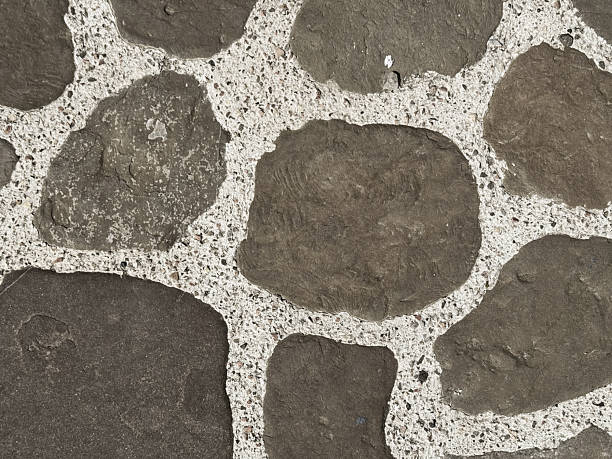 Stone mosaic pavement with natural dark rocks and light cement joints Close-up view of a textured stone wall featuring irregularly shaped stones set in a light-colored mortar. The stones vary in color and size, creating a rustic appearance.