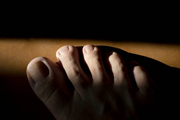 Partially illuminated toes rest on a brown surface, emerging from darkness into a strip of light, creating a dramatic contrast and highlighting the texture of the skin