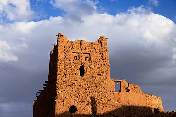 Ancient Moroccan kasbah made of mud bricks, showing signs of decay, stands tall against a backdrop of a cloudy sky in Tinghir