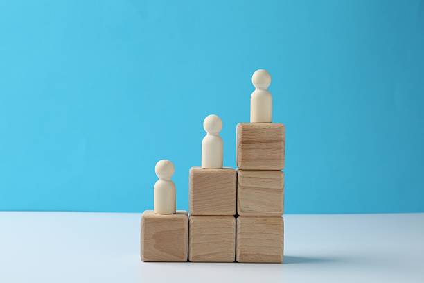 istock Human figures on stacked wooden blocks on white table against light blue background. Competition concept 2213221907