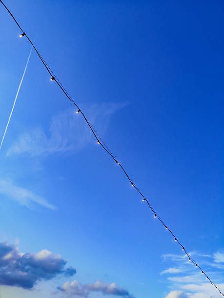 A string of decorative glowing light bulbs stretches across a clear blue sky, suggesting themes of celebration, outdoor lighting, or a festive atmosphere