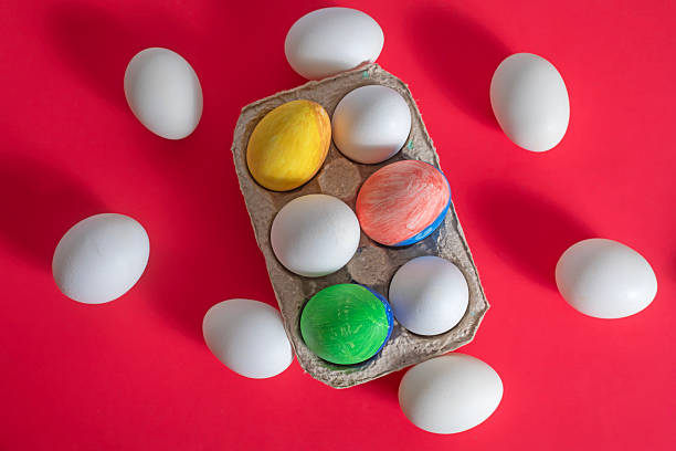 istock Overhead view of colored Easter eggs placed around white eggs in a cardboard box. 2213240684