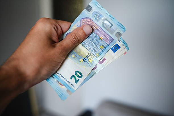 Close-up of a person's hand holding several euro banknotes, including 20 and 10 euro denominations, against a blurred indoor background. The composition highlights the colorful design and security features of the currency, evoking themes of finance, payment, and economic transactions.