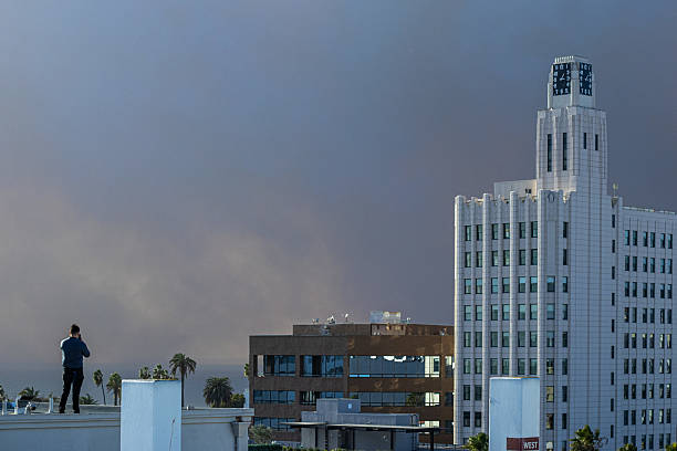 Los Angeles, California, USA. January 7th, 2025. The Pacific Palisades fire burns near Los Angeles, California. A man stands on a rooftop recording the scene on his smartphone as smoke drifts behind the Clock Tower Building in Santa Monica.