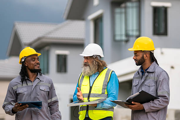 istock Three construction workers wearing hard hats and safety vests are standing in front of a house. They are holding papers and talking to each other 2213287917