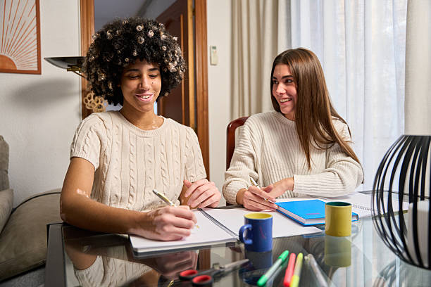 Two young women are sitting at a glass table, smiling and taking notes, studying together for an exam