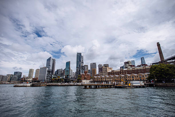 A waterfront view of Sydney's skyline showcasing modern skyscrapers alongside historic buildings under a cloudy sky, with a serene harbor in the foreground