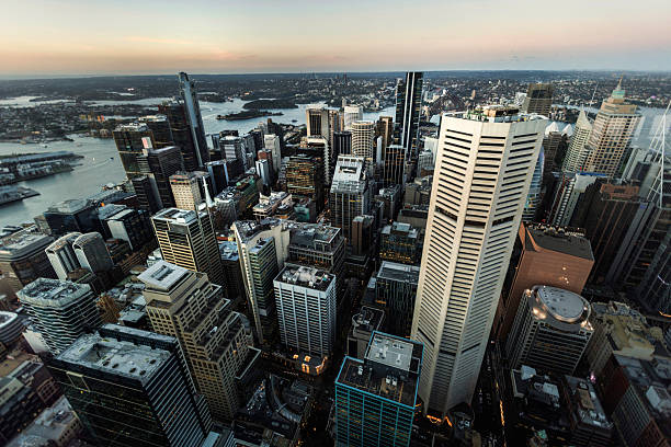An aerial view of Sydney's skyline during sunset, highlighting modern skyscrapers, the city's layout, and glimpses of the harbor under a soft twilight glow