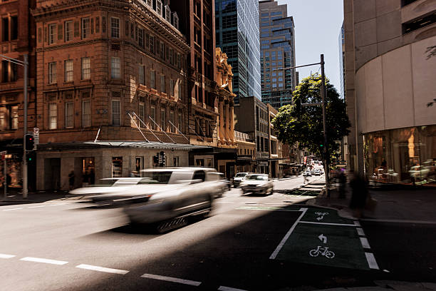 Vibrant Sydney street during daytime showcasing historic and modern architecture, sunlight filtering through buildings, and vehicles in motion; A dynamic urban view with a blend of activity and calm.