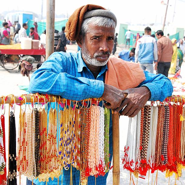 Street vendor selling beads chains and necklaces at the Prayagraj open air bazaar during the Kumbh Mehla festival. These days are unique to find a wide range of spiritual memorabilia and religious trinkets. \nThe Maha Kumbh Mela  also provides  an economic opportunity for many  workers and small vendors who are able to utilise these 45 days religious gathering as a source of livelihood by selling all sort of essentials and services.