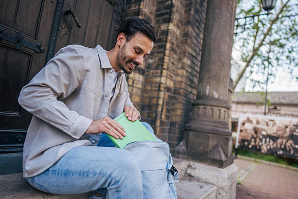 Young adult male student sitting on steps outside a university building, putting a book into his backpack while enjoying a cheerful moment in the vibrant campus atmosphere