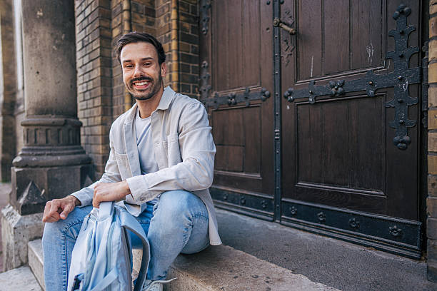 Portrait of a cheerful university student sitting on the steps in front of a university building, holding his backpack and smiling at the camera