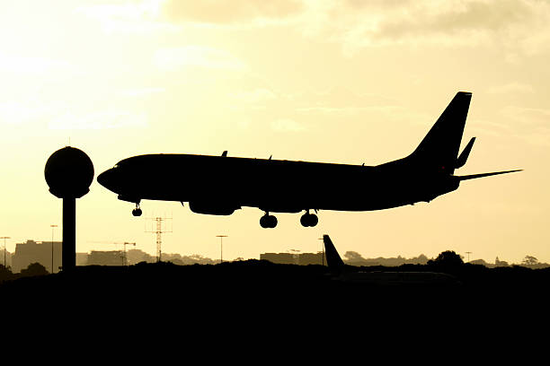 An Air Niugini Boeing B737-86Q plane, registration P2-PXC, landing on the third runway of Sydney Kingsford Smith Airport as flight PX1 from Port Moresby. \n\nIn the left background is the airport's weather measuring installation.  In the right background is the vertical stabiliser of a Virgin Australia Boeing B737-8FE plane, registration VH-YVC, taxiing for departure as fight VA533 to the Gold Coast. \n\nThis image faces west and was taken from Mill Stream Lookout, Botany Bay on a cold and stormy afternoon approaching sunset on 2 May 2025.