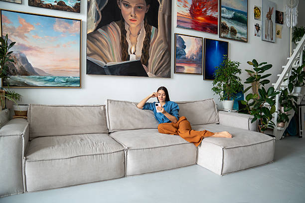 istock Peaceful woman using phone, lying relaxing on sofa in home art studio among paintings and plants. 2213326495