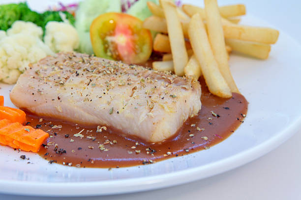 Grilled Pangasius Fillet, Delicious grilled fish steak served with pepper sauce, French fries, steamed vegetables, and fresh salad on a white plate, perfect for healthy meal concepts