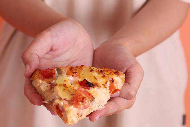 Hand holding a slice of Hawaiian pizza, a pizza topped with tomato sauce, cheese, pineapple, and ham or bacon on a white plate, isolated on an orange background. Italian food, Italian cuisine.