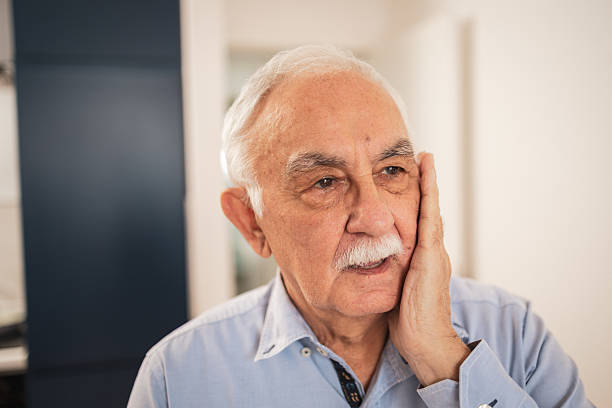 Elderly man holding his cheek with a concerned expression indoors