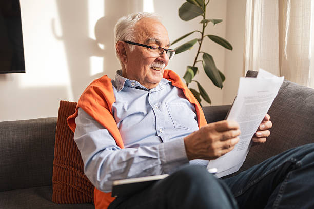 Senior man reading documents while sitting on a sofa in bright sunlight An elderly man in glasses and casual clothing attentively reviews papers while relaxing at home, bathed in natural light. The setting feels thoughtful, comfortable, and focused. happy old man  buying life insurance stock pictures, royalty-free photos & images
