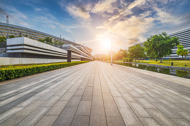 Empty stone pavement pathway leading towards modern buildings with green landscaping under a sunset sky in city.