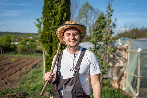 A happy male farmer wearing a straw hat and standing in a well-maintained garden holding a wooden tool. The lush greenery and clear sky create a serene and productive ambiance.