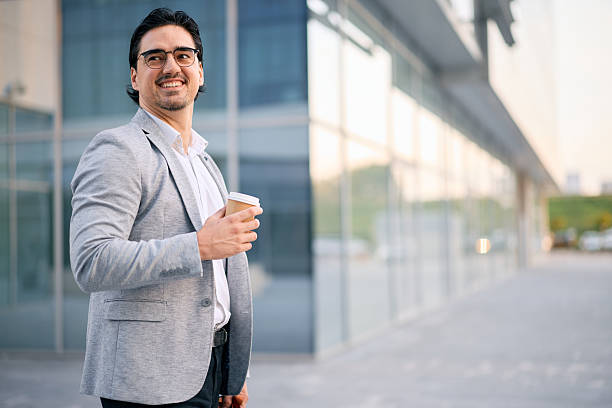 istock Smiling businessman walking and drinking coffee outside modern office building 2213370605
