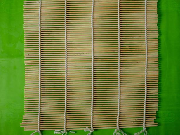 Flat lay image of a traditional bamboo sushi mat placed on a vivid green background. The empty mat sets a clean and organized scene for the beginning of Japanese food preparation, often used in homemade sushi making. This minimal composition focuses on texture, symmetry, and culinary context, making it suitable for instructional materials, recipe visuals, or food blog illustrations. Ideal for showcasing kitchen tools, step-by-step guides, or Asian cuisine culture, the photo captures a natural and clean setup perfect for visual storytelling.