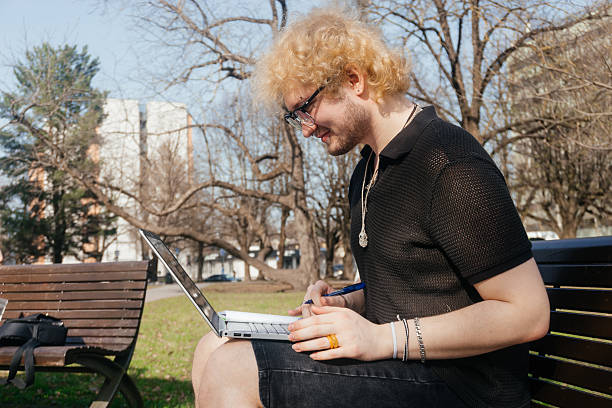 A focused student works outdoors on a laptop and takes notes while sitting alone on a park bench on a sunny day